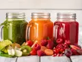 Trzy słoiki z kolorowymi sokami: zielonym, pomarańczowym i czerwonym. Obok owoce i warzywa, idealne na dietę sokową.