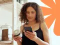 Kobieta sprawdza na telefonie, czy produkt ma dużo węglowodanów. Na półce stoją słoje z produktami.