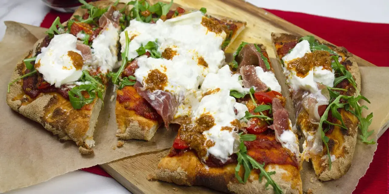 Pyszna pinsa z burratą, rukolą i szynką parmeńską, podana na desce.