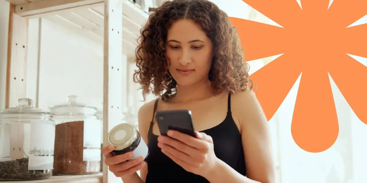 Kobieta sprawdza na telefonie, czy produkt ma dużo węglowodanów. Na półce stoją słoje z produktami.