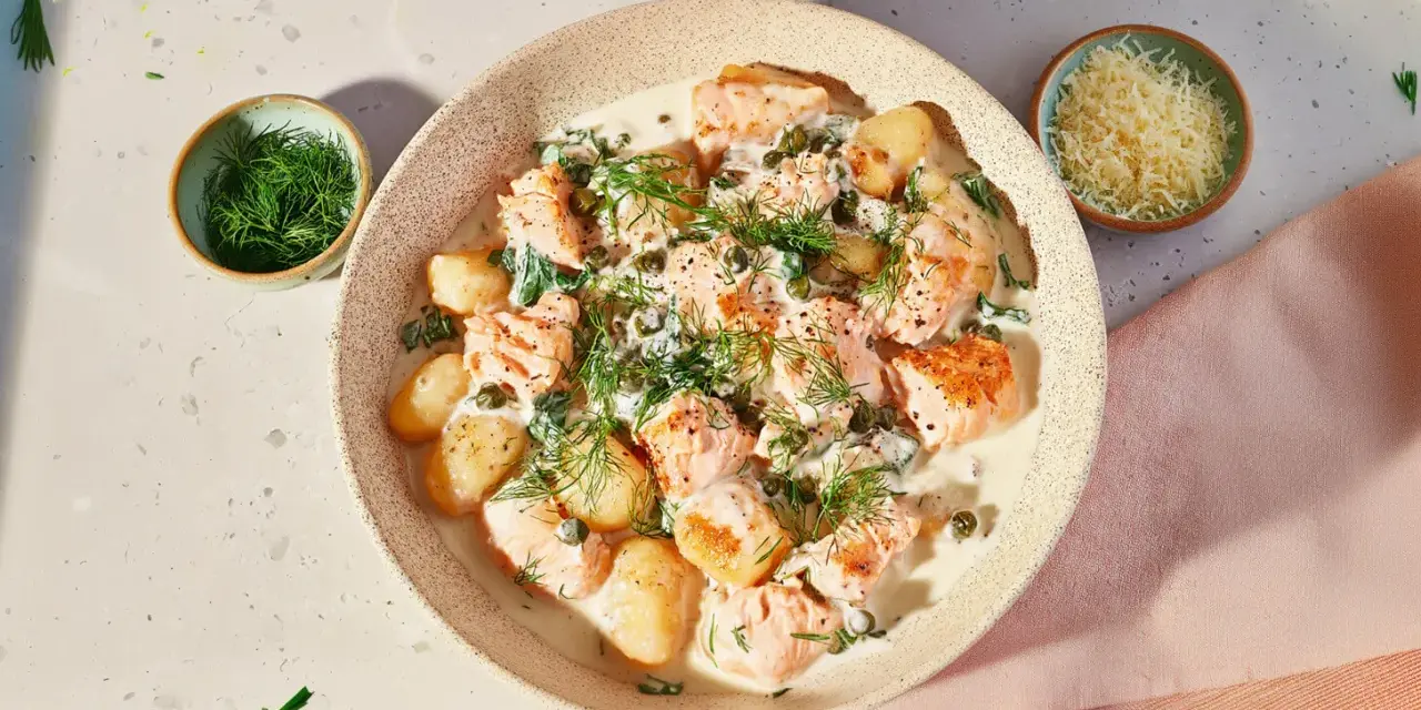 Pyszne gnocchi z łososiem w kremowym sosie, udekorowane koperkiem i kaparami. Obok miseczka z tartym serem.