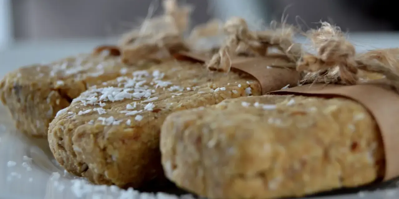Domowe batony proteinowe przepis. Trzy domowe batony proteinowe, posypane wiórkami kokosowymi, zawinięte w papier i związane sznurkiem.