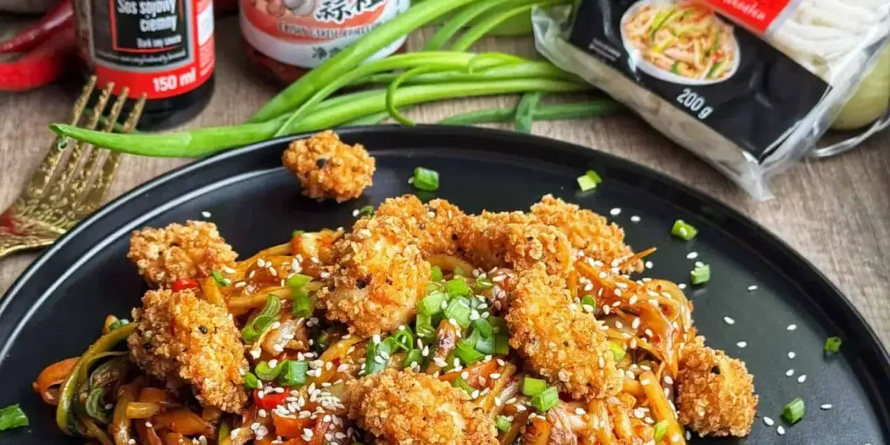 Pyszny makaron udon z kurczakiem w panierce, posypany sezamem i szczypiorkiem. Obok sosy i opakowanie makaronu.