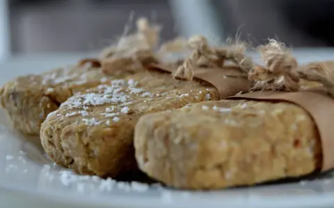 Domowe batony proteinowe przepis. Trzy domowe batony proteinowe, posypane wiórkami kokosowymi, zawinięte w papier i związane sznurkiem.