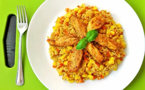 Pyszna kasza bulgur z kurczakiem, kukurydzą i marchewką, udekorowana listkiem bazylii. Obok leży widelec.