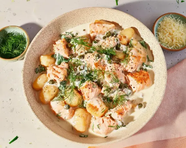 Pyszne gnocchi z łososiem w kremowym sosie, udekorowane koperkiem i kaparami. Obok miseczka z tartym serem.