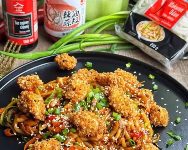 Pyszny makaron udon z kurczakiem w panierce, posypany sezamem i szczypiorkiem. Obok sosy i opakowanie makaronu.