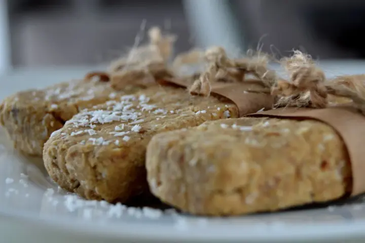 Domowe batony proteinowe przepis. Trzy domowe batony proteinowe, posypane wiórkami kokosowymi, zawinięte w papier i związane sznurkiem.