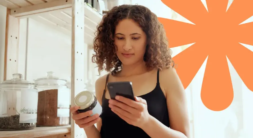 Kobieta sprawdza na telefonie, czy produkt ma dużo węglowodanów. Na półce stoją słoje z produktami.