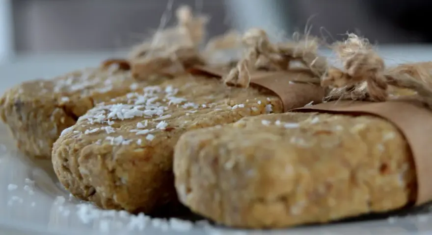 Domowe batony proteinowe przepis. Trzy domowe batony proteinowe, posypane wiórkami kokosowymi, zawinięte w papier i związane sznurkiem.