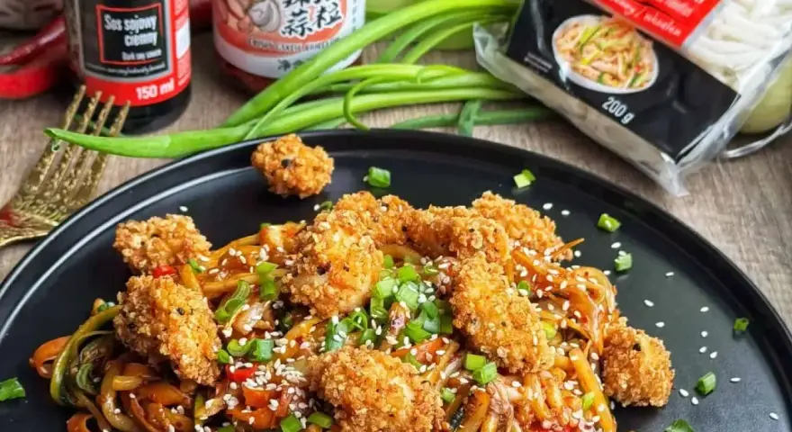 Pyszny makaron udon z kurczakiem w panierce, posypany sezamem i szczypiorkiem. Obok sosy i opakowanie makaronu.