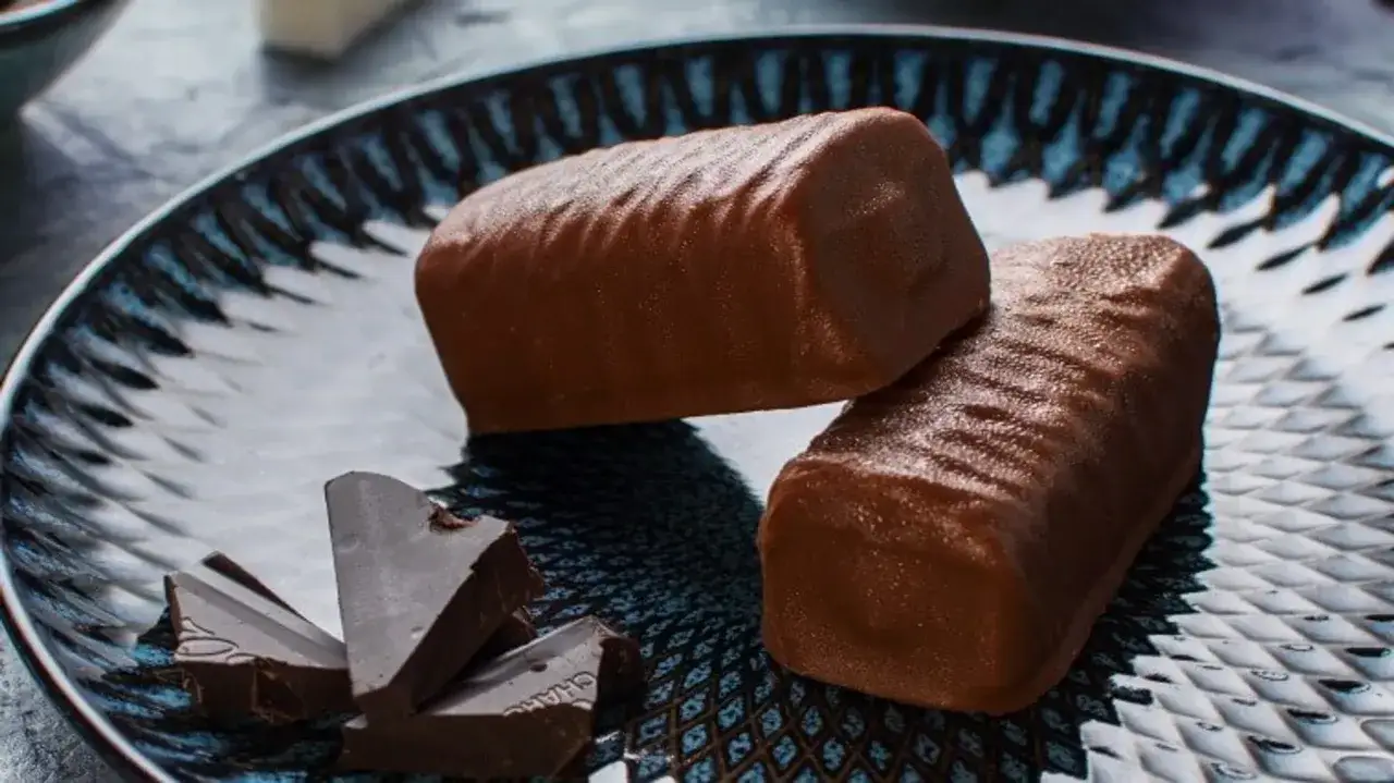 Dwa czekoladowe batony proteinowe na talerzu, obok kawałki gorzkiej czekolady. Idealne na zdrową przekąskę, gdy szukasz przepisu na batony proteinowe.