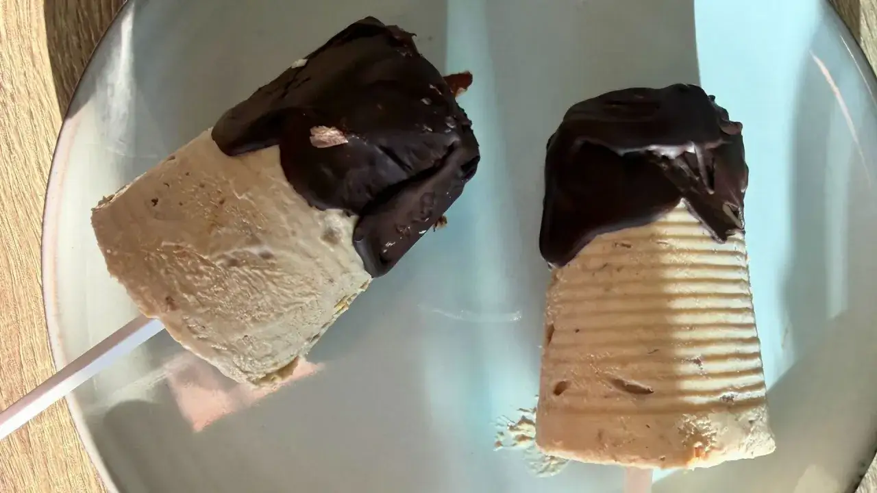 Dwa lody na patyku z serka wiejskiego, oblane czekoladą, leżą na talerzu.