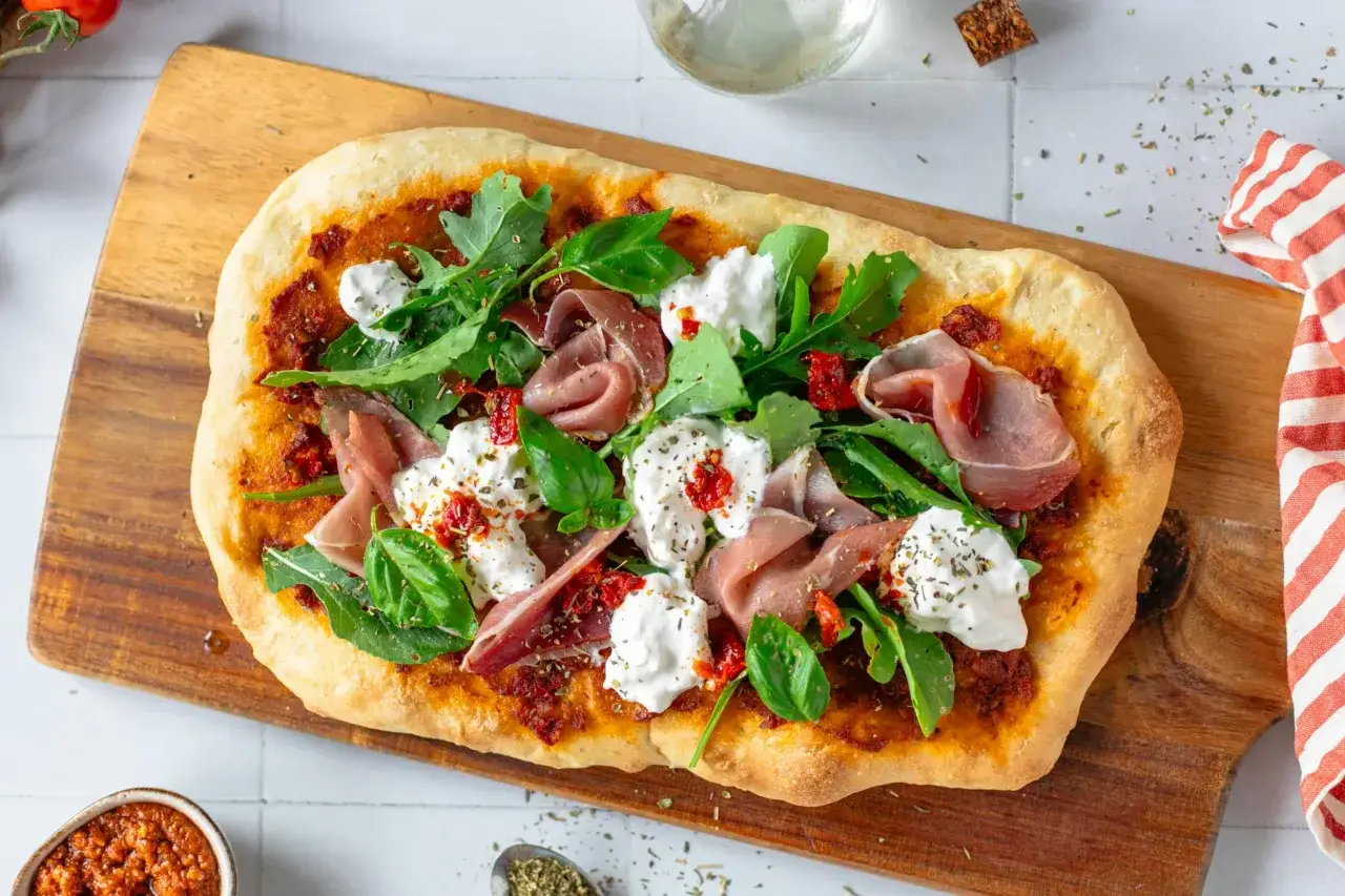 Pyszna pinsa z burratą, rukolą i szynką parmeńską, ozdobiona suszonymi pomidorami i ziołami.