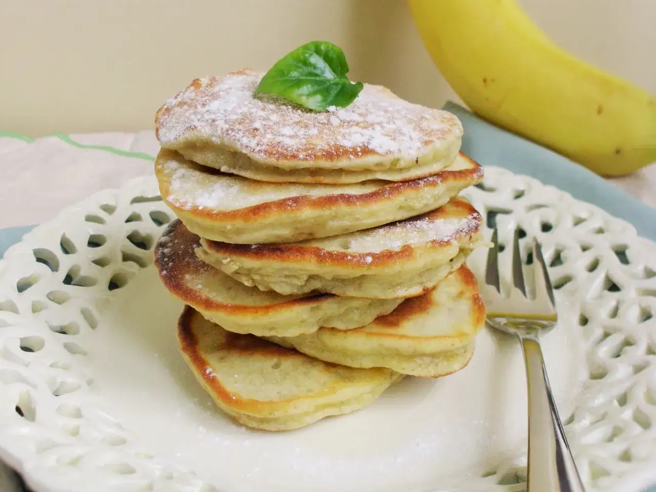 Stos puszystych placuszk&oacute;w bananowych, posypanych cukrem pudrem, z listkiem mięty na wierzchu. Obok leży banan.